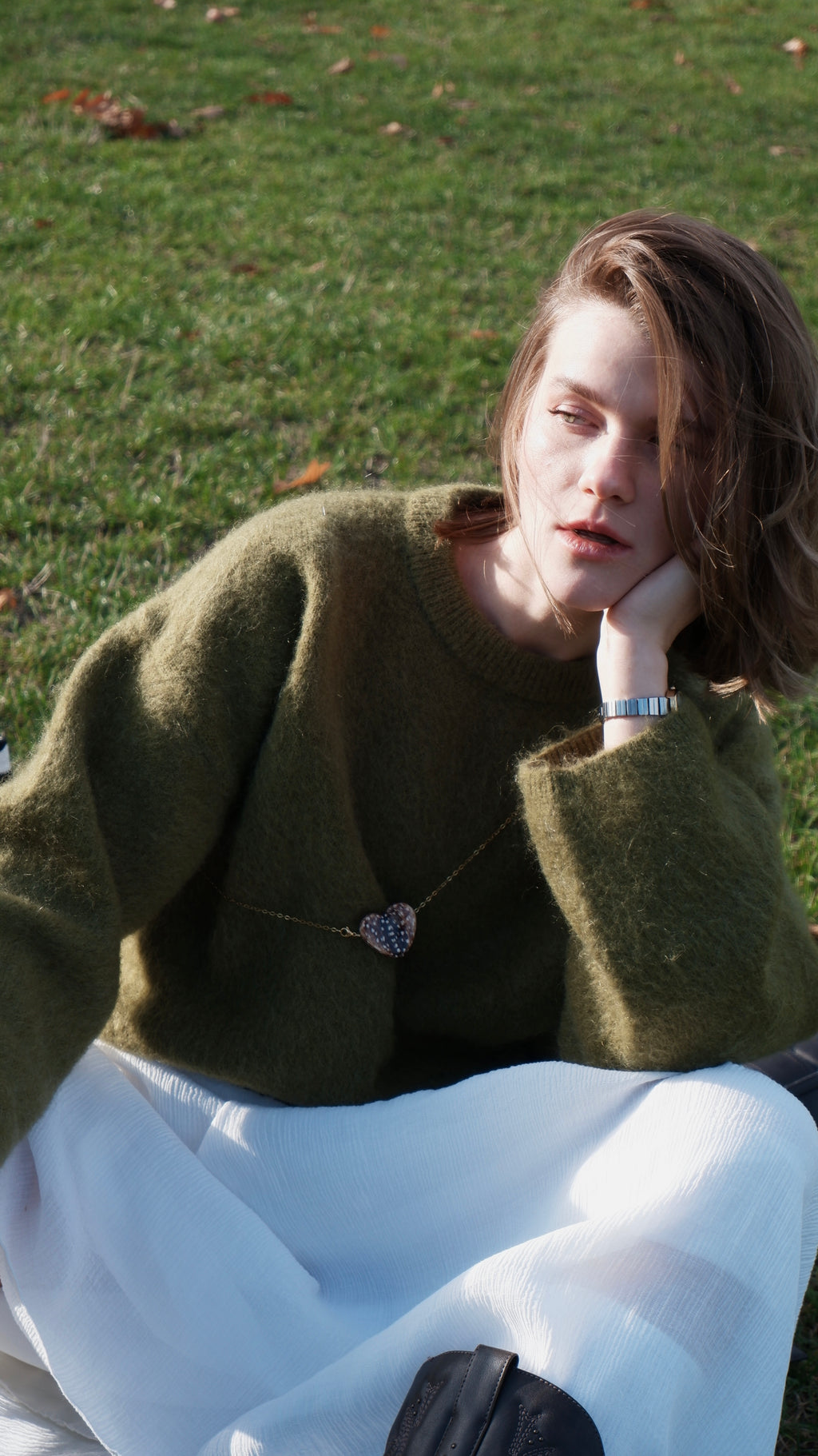 Girl wearing a pedant in heart shape and baby deer print in green coat and white dress sitting on grass